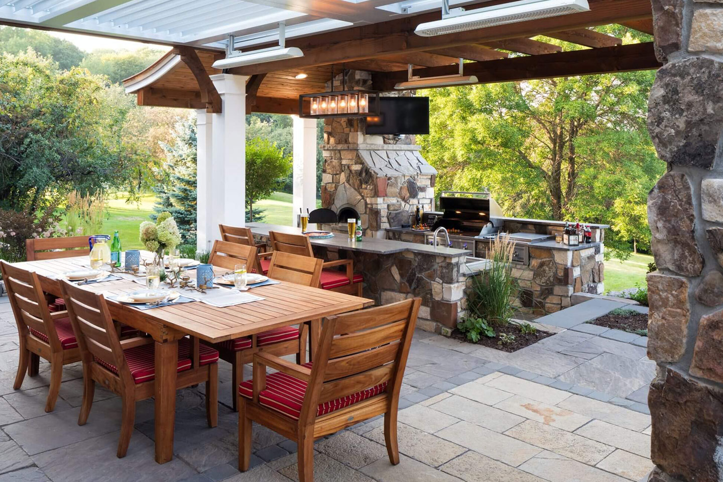 Custom outdoor kitchen design with a rustic stone island, built-in stainless steel grill, and large wooden dining table under a protective pergola, perfect for backyard entertaining and premium home improvement.