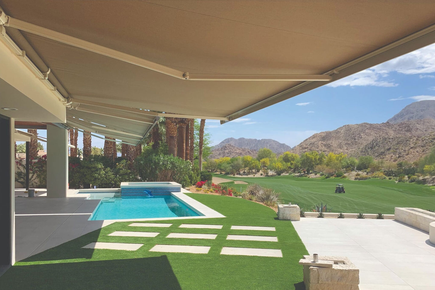 Retractable patio awning by Decadent Outdoors installed over a luxury poolside deck and turf lawn, providing custom shade for a backyard overlooking a lush green golf course and mountain landscape.
