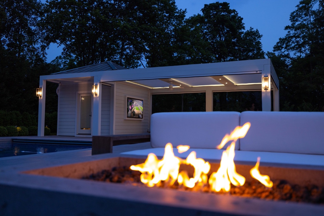A close up of a fireplace with a StruXure pergola in the background in the evening hours, complete with lighting, creating the perfect atmosphere.